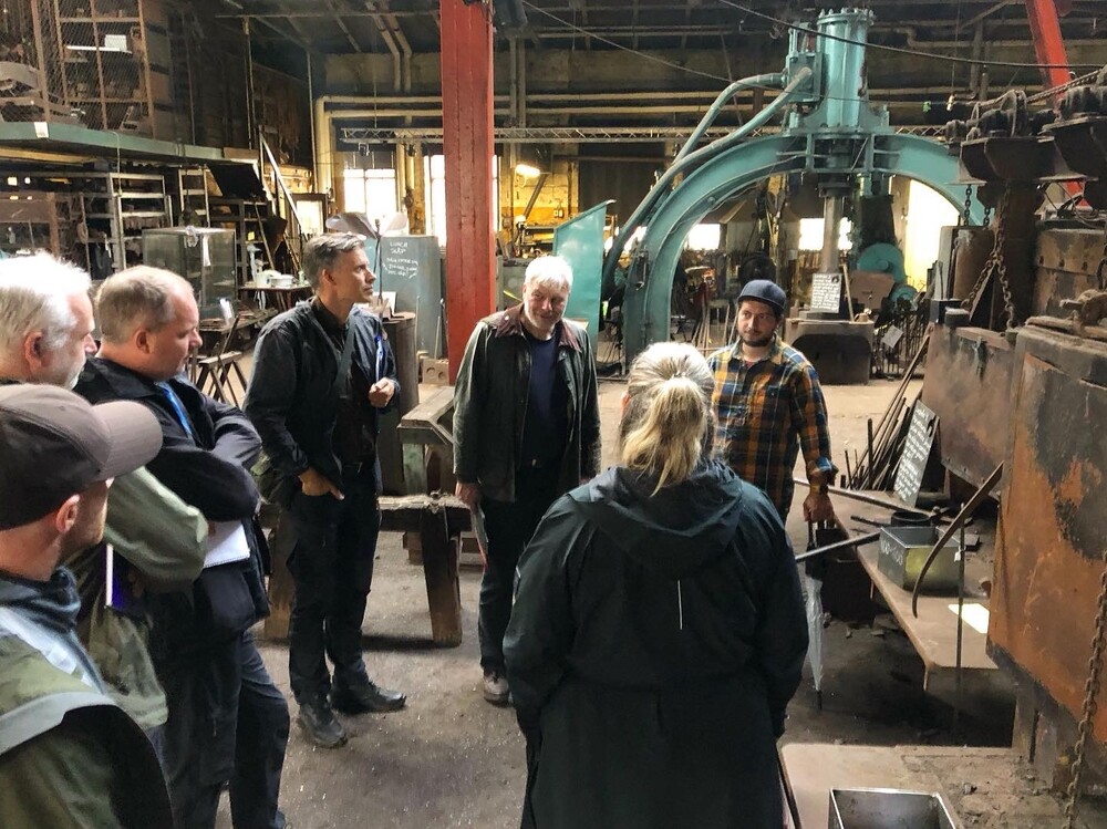 En grupp människor står och lyssnar på en guide i en industrilokal med maskiner och verktyg.