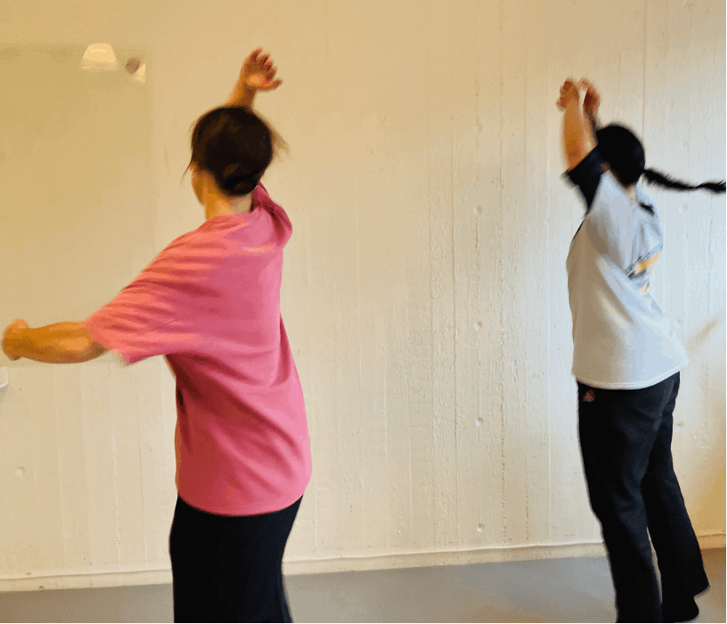 Två personer dansar eller tränar med armarna uppsträckta i ett ljust rum med vit vägg.