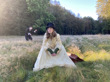 En person i gammaldags klänning sitter på en äng, med en annan person i bakgrunden och skog runtom.