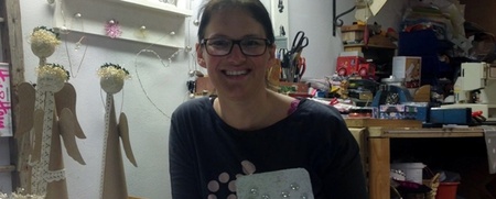 Kvinna med glasögon sitter vid ett bord och visar upp ett handgjort konstverk i en pysselverkstad.