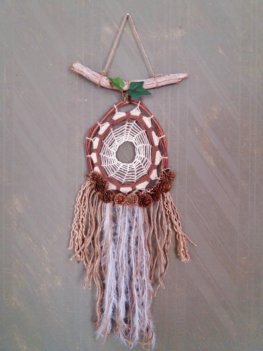 En handgjord drömfångare med naturmaterial, hängande mot en grå vägg.