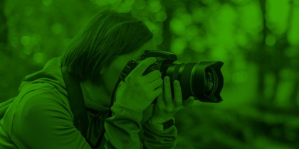 Person fotograferar i skogen med en kamera, bilden har ett grönt filter.