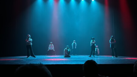 Sju personer står och sitter på en scen med blå och röd belysning, redo för dansuppträdande.