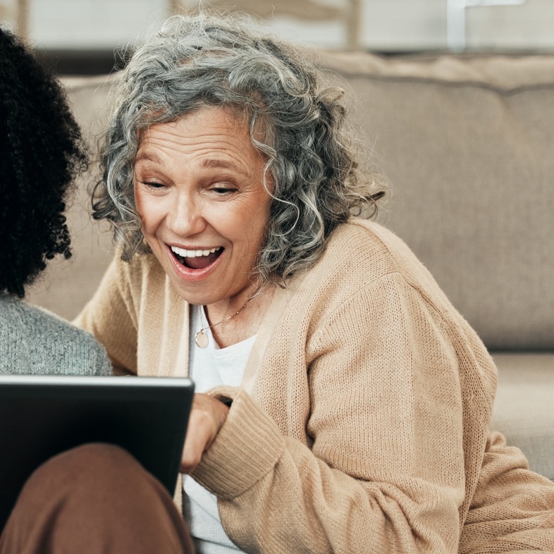 Två kvinnor sitter på golvet framför en soffa och tittar glatt på en surfplatta tillsammans.