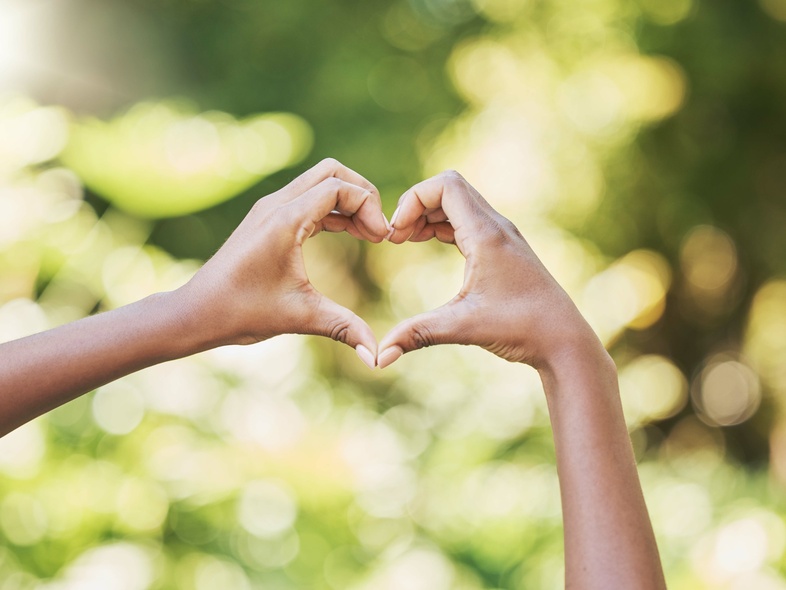 Två händer formar ett hjärta mot en suddig, grön bakgrund utomhus.
