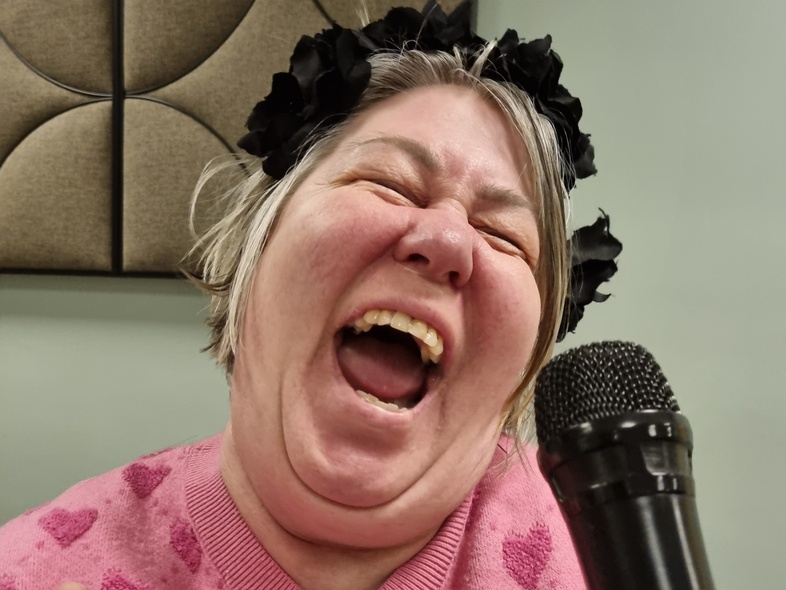 Kvinna i rosa tröja med hjärtan skrattar och håller en mikrofon, glasögon hänger runt halsen.