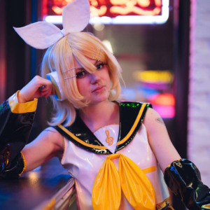 Person i cosplay med blont hår, stora vita rosetter och gul-svart sjömansdräkt poserar inomhus vid ett bord.
