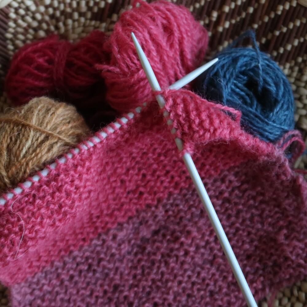 Närbild på stickning med rosa garn, stickor och garnnystan i olika färger.