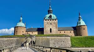Stort slott med tre gröna tornkupoler, blå himmel och personer som går på en bro mot ingången.