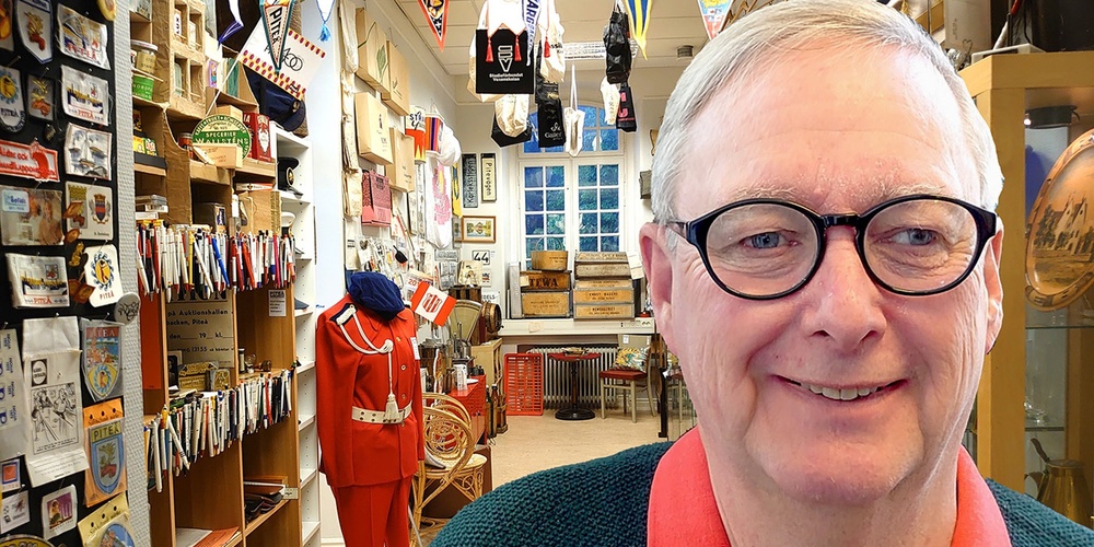 Äldre man med glasögon ler i ett rum fullt av samlarföremål och memorabilia.