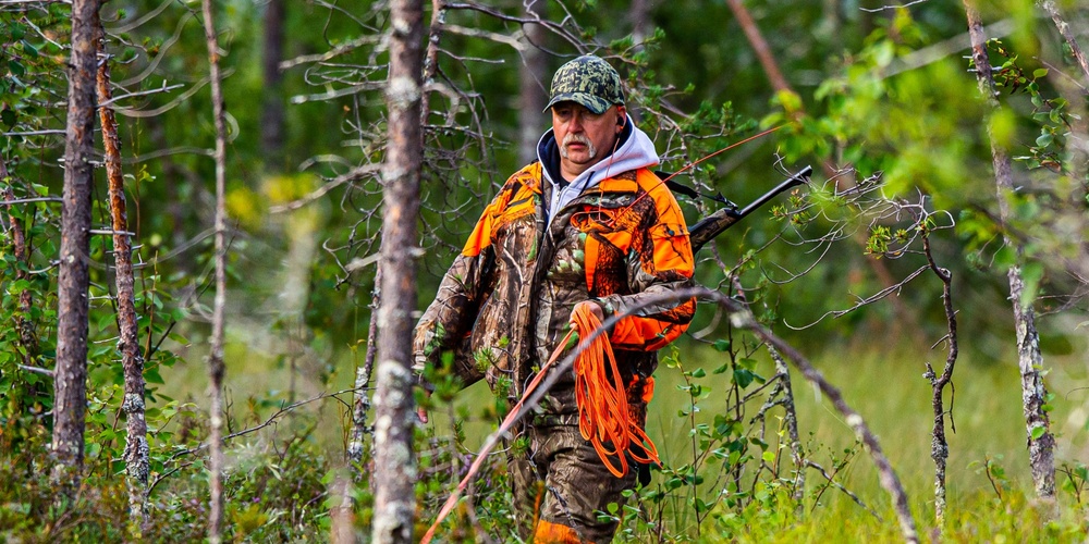 En jägare i orange och kamouflagekläder går med gevär genom en grön skog.