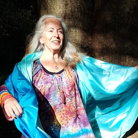 Cirkelledare Elisabeth Hjulström med långt grått hår står barfota utomhus, bär färgglad tunika och blå sjal.