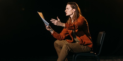 En person sitter på en stol på en scen och gestikulerar med ett manus i handen under en teaterövning.