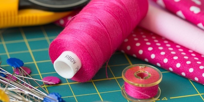 Närbild av sybehör: rosa tråd, knappnålar, rullkniv och rosa tyg med vita prickar och hjärtan.