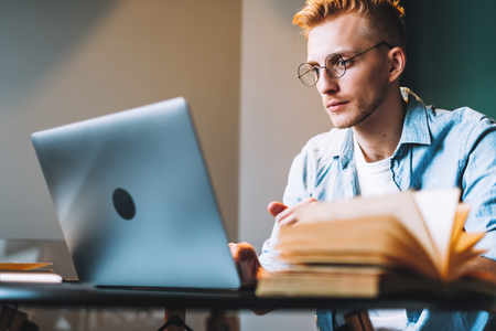 Ung man med glasögon studerar vid en bärbar dator och har en uppslagen bok framför sig.