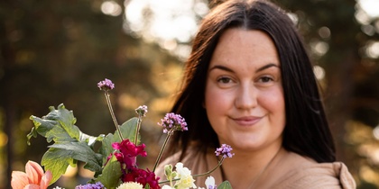 Person håller en stor bukett färgglada blommor utomhus i naturligt ljus.