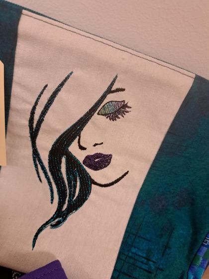 Broderad bild av ett stiliserat kvinnligt ansikte med lila läppar och grönt öga på tyg.