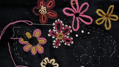 Broderade blommor i rosa, gult och brunt på svart tyg, med nål och tråd synliga.
