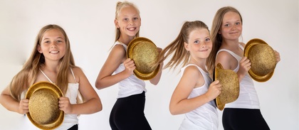 Fyra unga tjejer i vita linnen och svarta byxor poserar med glittriga guldhattar mot en ljus bakgrund.