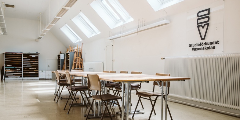 Ljust klassrum med tomma stolar och bord, stora fönster och skylten 'Studieförbundet Vuxenskolan' på väggen.