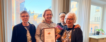 Fyra kvinnor står tillsammans inomhus, en håller ett diplom och blommor, alla ser glada ut.