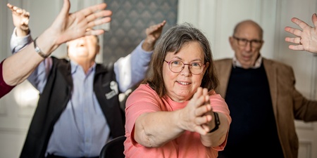 En grupp äldre personer gör gymnastik tillsammans inomhus, en kvinna i rosa tröja i fokus.