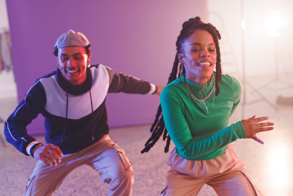 Två unga personer dansar energiskt i ett färgglatt rum med lila bakgrund.