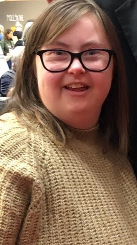 Person i beige stickad tröja sitter inomhus bland andra människor.