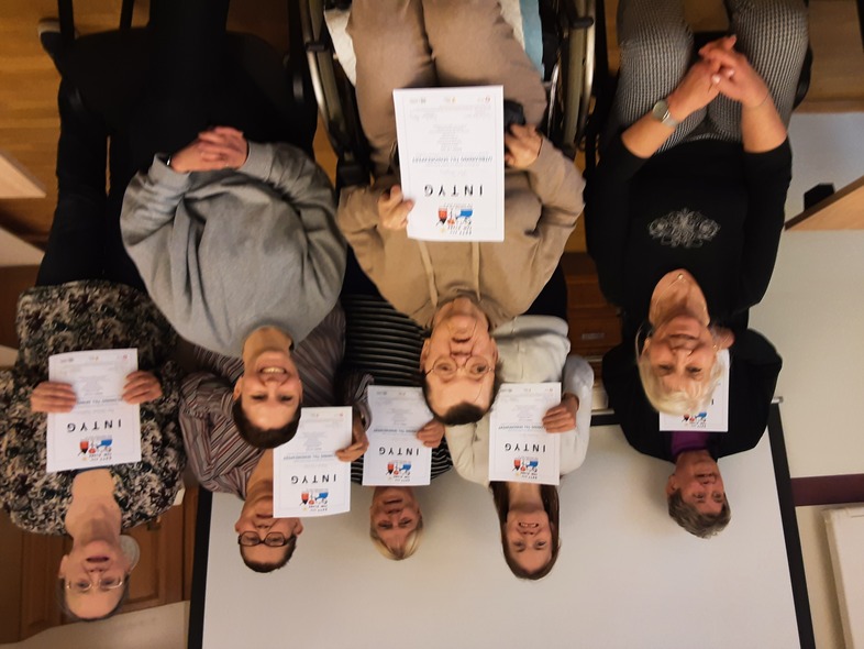 Gruppfoto av åtta personer, flera håller upp intyg och ler mot kameran i ett inomhusrum.