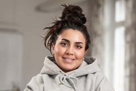 Kvinna med mörkt hår i knut och grå hoodie står med armarna i kors i ett klassrum.