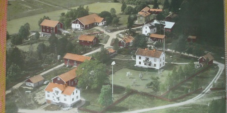 Flygfoto över en liten by med röda och vita hus, omgiven av grönska och åkrar.