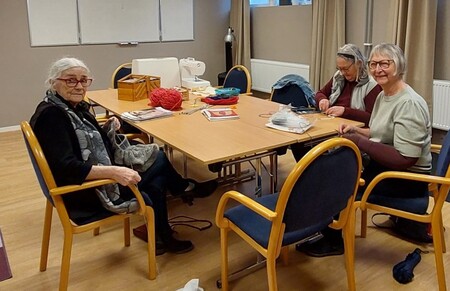 Tre personer sitter runt ett bord och handarbetar i ett ljust rum med trästolar och handarbetsmaterial.