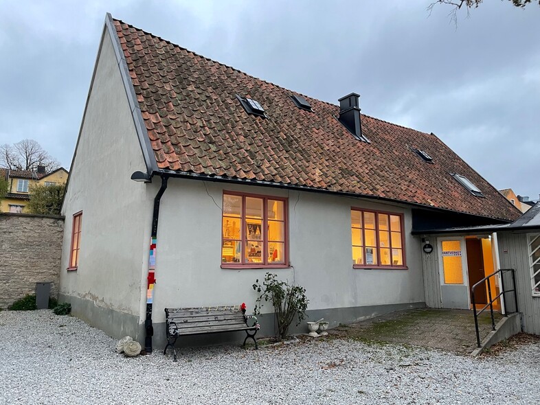 Ett litet hus med tegeltak och grå fasad, upplysta fönster och en bänk utanför, på en grusad gård.