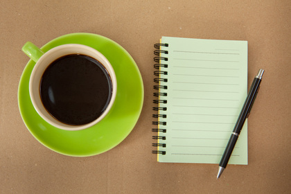 En kopp svart kaffe på ett grönt fat bredvid ett tomt spiralblock och en penna på ett brunt bord.
