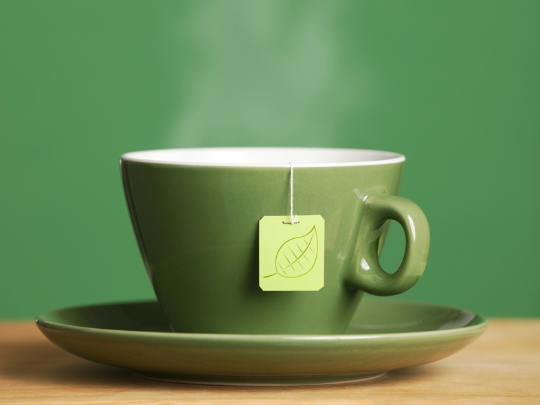 En grön kopp med te på ett fat, med en tepåse och ånga som stiger upp, mot grön bakgrund.