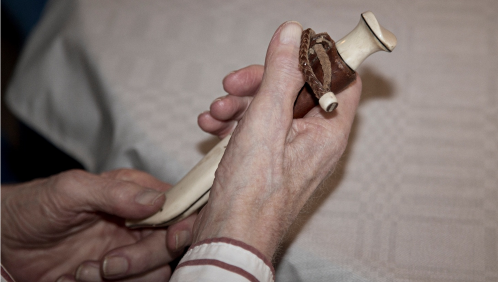 Två händer håller en liten dekorativ kniv med slida, liggande på ett bord med vit duk.