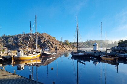 Små segelbåtar ligger förtöjda vid en brygga i en lugn hamn med klippor och blå himmel i bakgrunden.