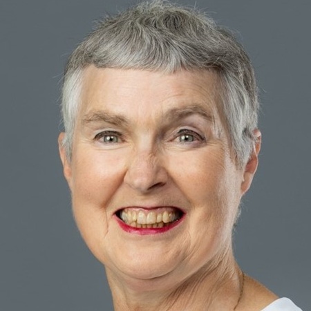 Äldre kvinna med kort grått hår och vit skjorta ler mot kameran mot grå bakgrund.