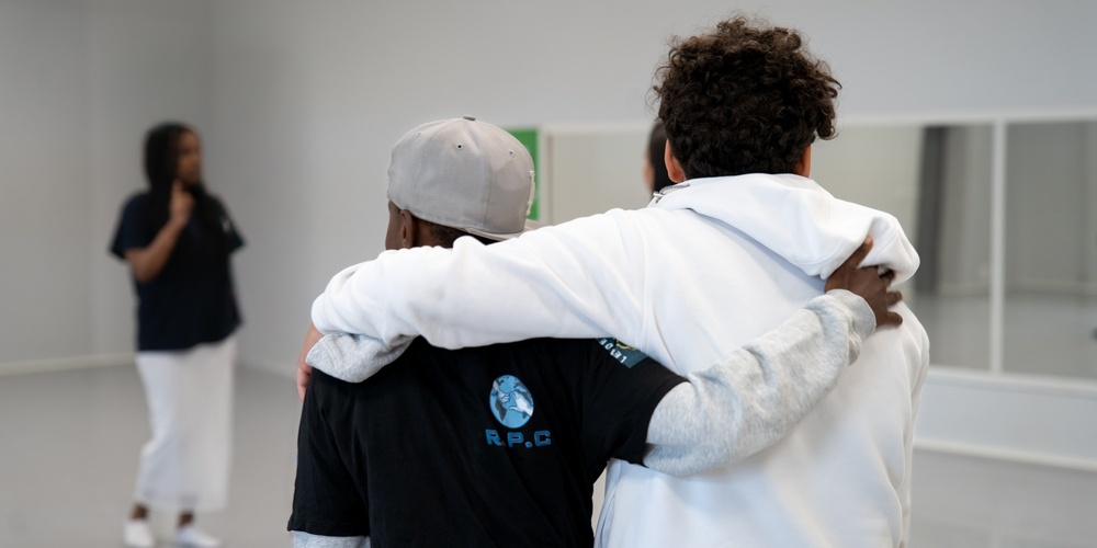 Två personer står med armarna om varandra i ett ljust rum, en tredje person syns i bakgrunden.