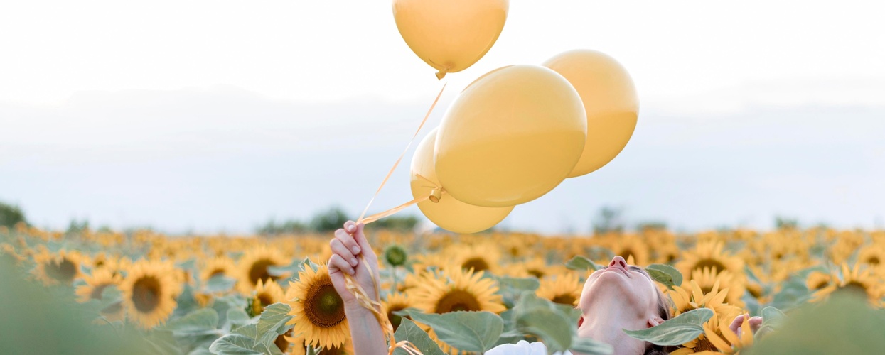 Person håller tre gula ballonger i ett fält av solrosor under en ljus himmel.