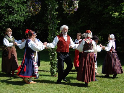 En grupp människor i folkdräkt dansar runt en midsommarstång utomhus i solskenet.