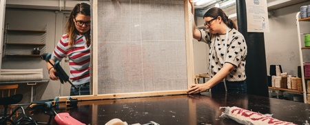 Två personer arbetar tillsammans med en stor träram i en verkstad med textilmaterial.