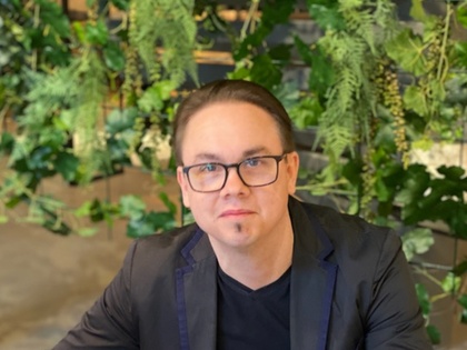 Person i svart kavaj sitter vid ett bord och bläddrar i en tidning, omgiven av gröna växter.