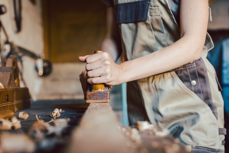 En person i arbetskläder hyvlar trä i en snickarverkstad, fokus på händer och verktyg.