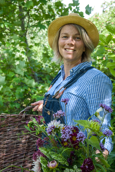 Kvinna med halmhatt och blå skjorta håller en korg med blommor i en grönskande trädgård.