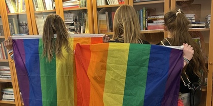 Tre personer står med ryggen mot kameran och håller upp en regnbågsflagga framför en bokhylla.