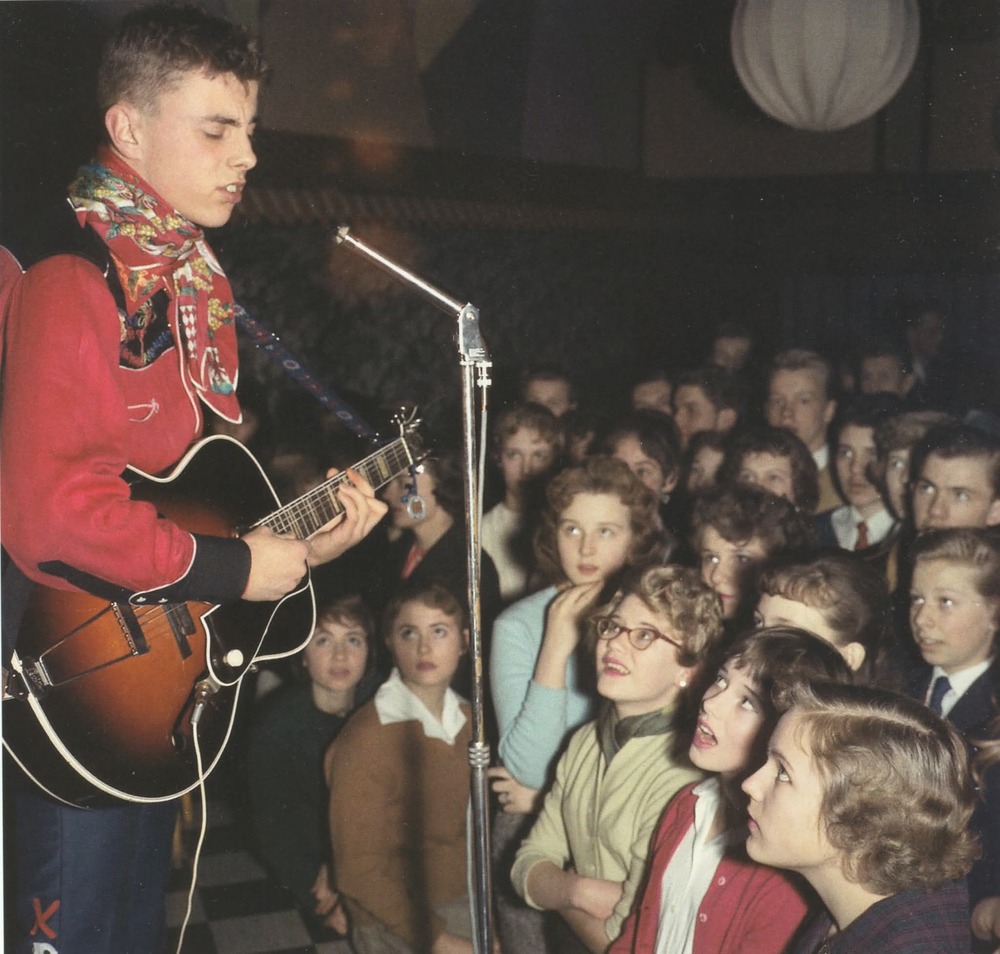 Ung man spelar gitarr och sjunger på scen inför en entusiastisk publik av ungdomar.