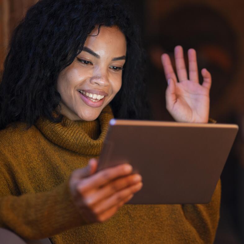 Kvinna med lockigt hår vinkar och ler mot en surfplatta i ett varmt upplyst rum.