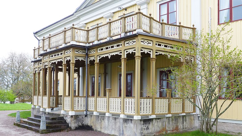 Gul trävilla med snickarglädje och stor veranda, omgiven av grönska.