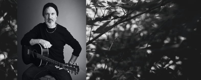 Man med skägg och mössa sitter med en akustisk gitarr, svartvit bild, naturmotiv till höger.